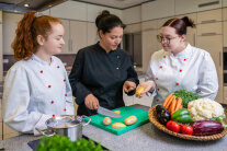 3 Personen stehen nebeneinander in einer Küche, vor ihnen Schale mit Gemüse 